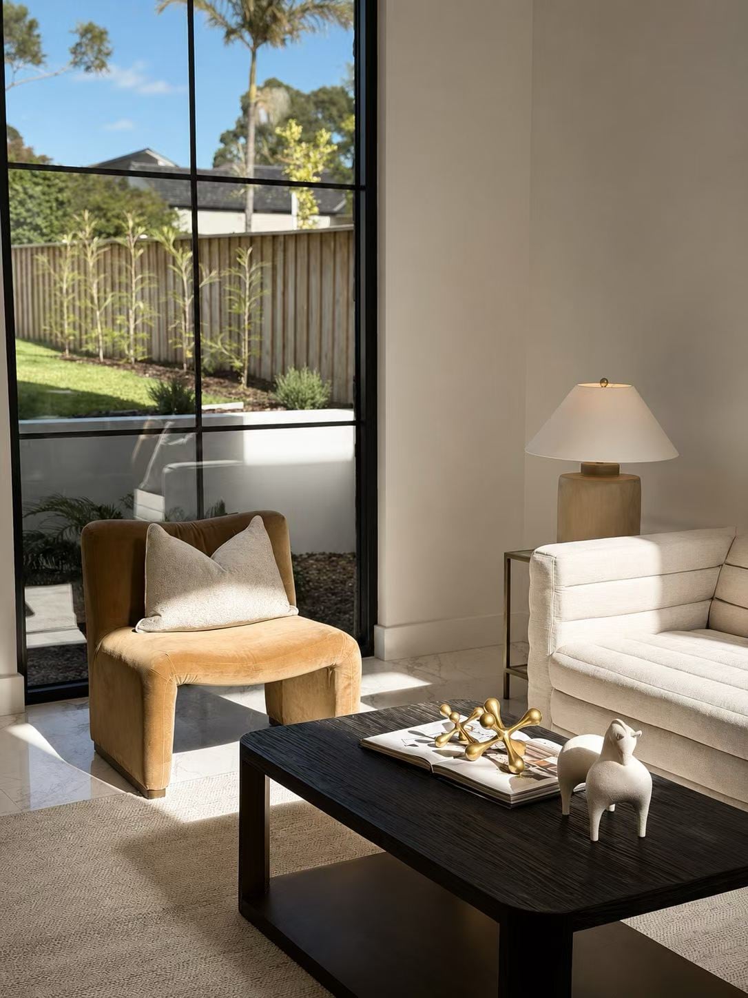 Tan bouclé armchair and a black coffee table beside a tall window to the garden, with a cream sofa and table lamp