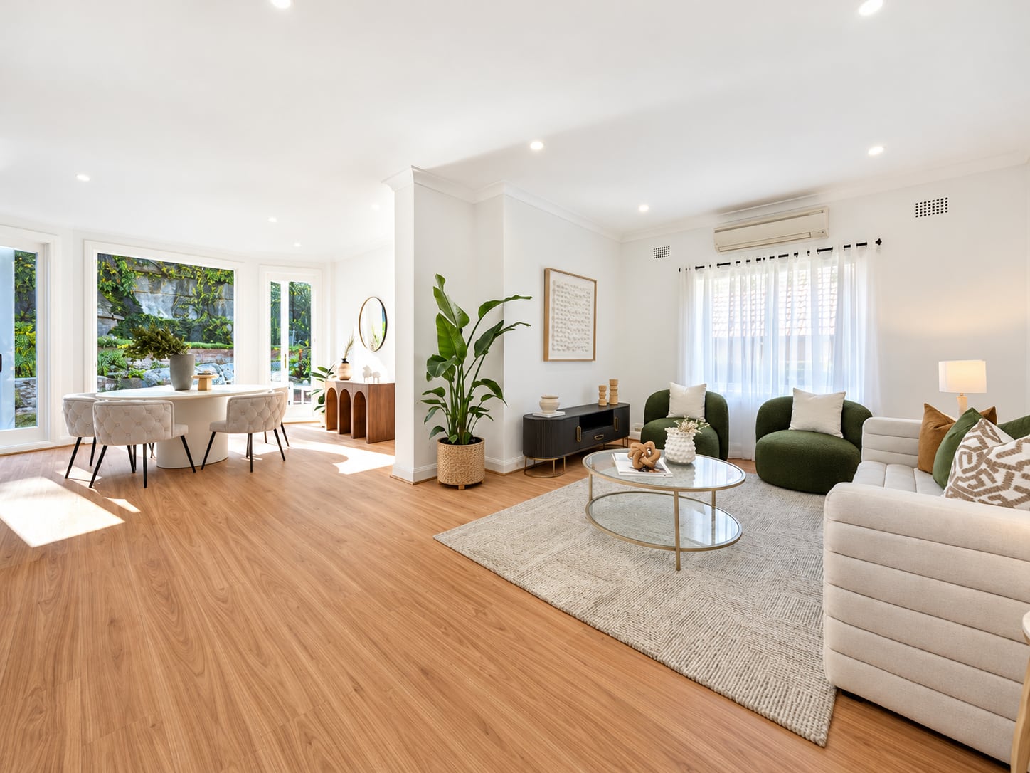 Open-plan living and dining room with a channelled cream sofa, green bouclé armchairs, a glass coffee table and a round white dining table by the garden windows