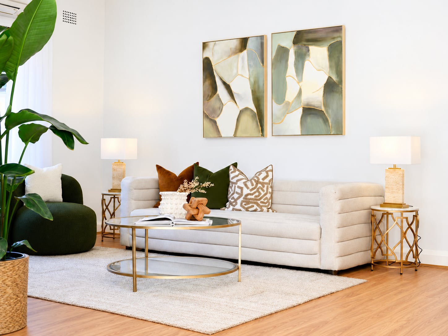 Channelled cream sofa with rust, green and patterned cushions beneath two framed abstract artworks, between gold side tables and stone lamps
