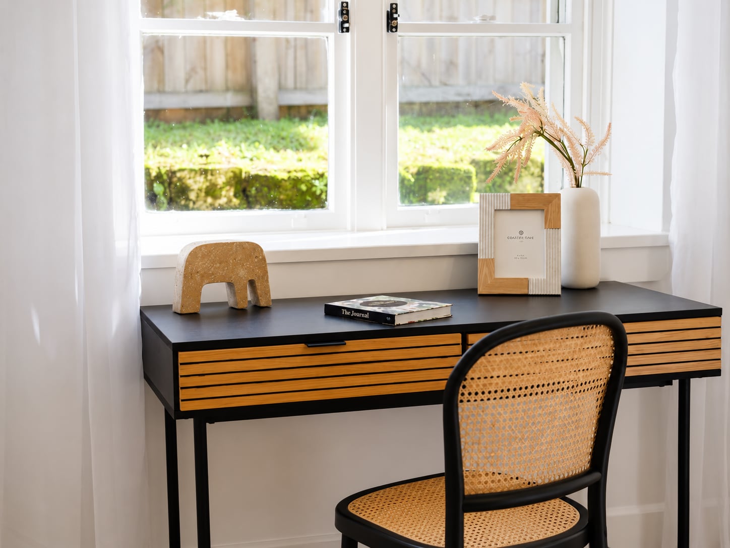 Black study desk with timber-slatted drawers and a rattan chair beneath a garden window