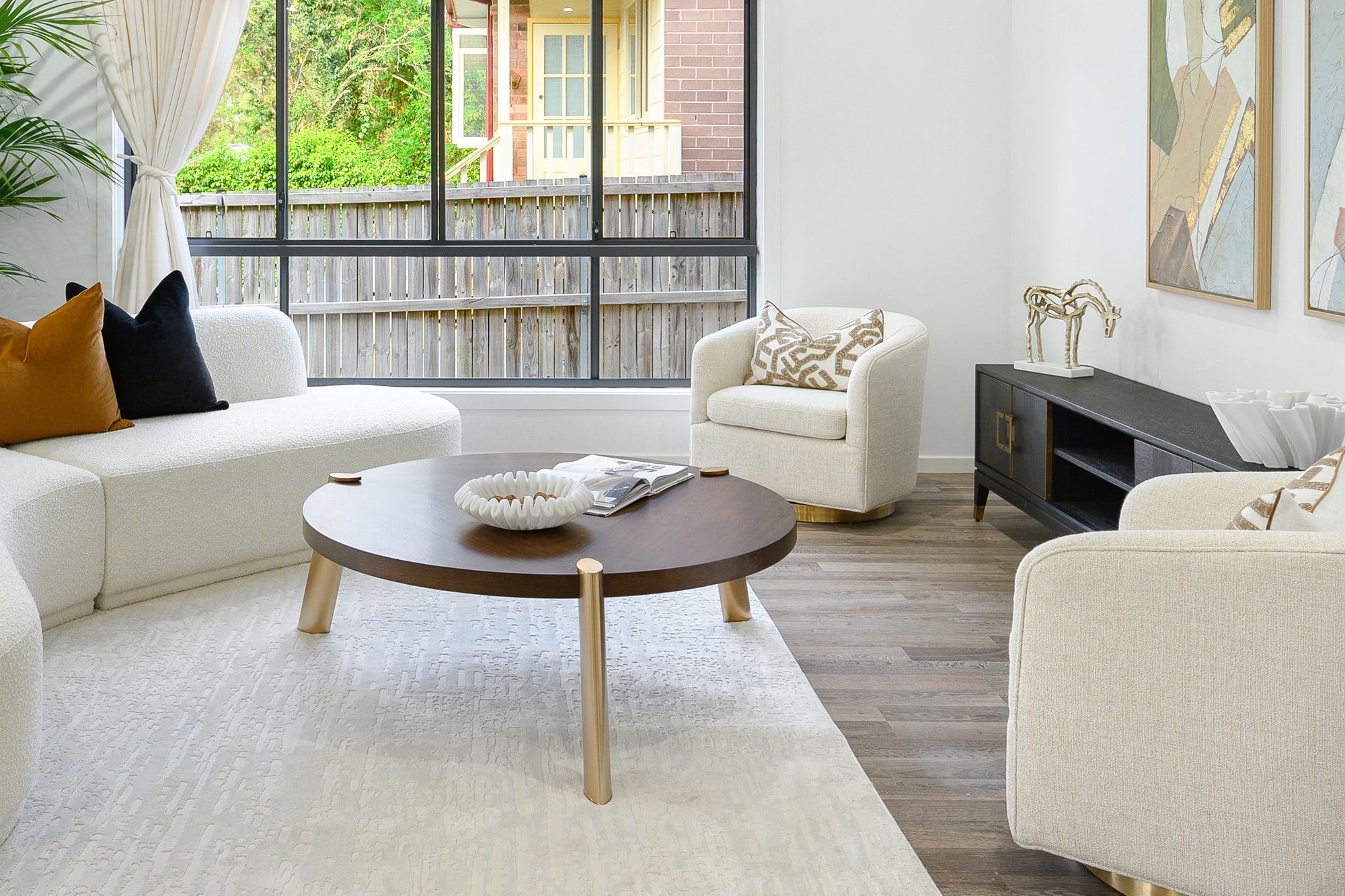 Round timber coffee table with gold legs in front of a cream armchair and a framed abstract artwork