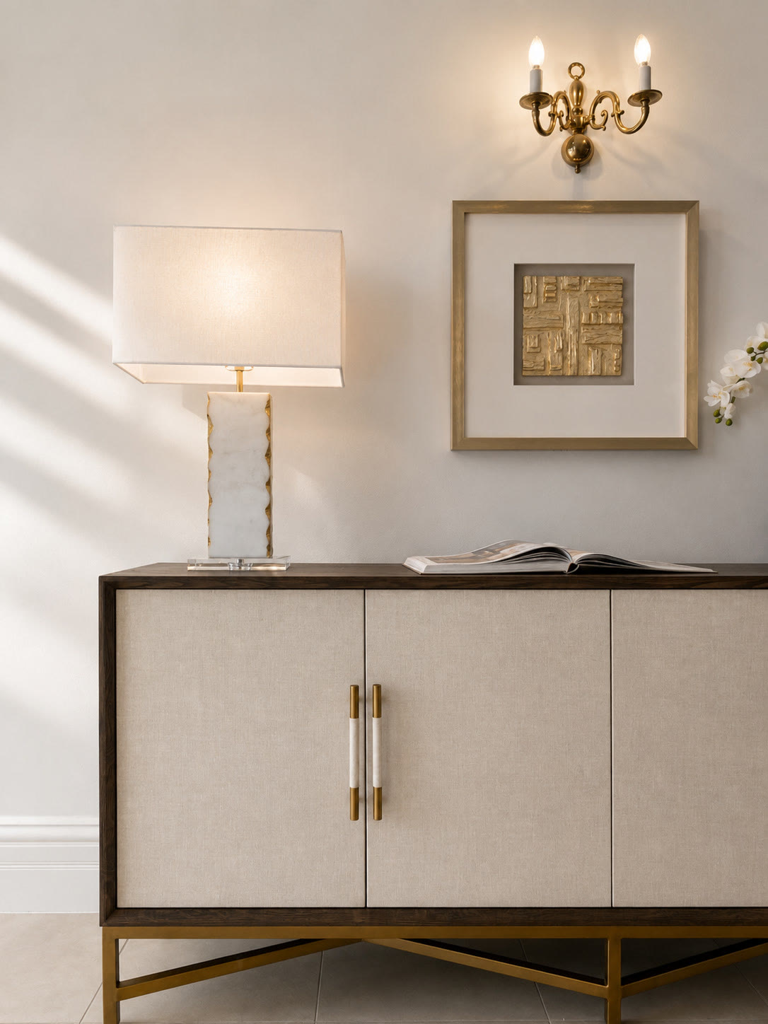 Cream sideboard with brass handles, a lamp and framed gold artwork under a wall light