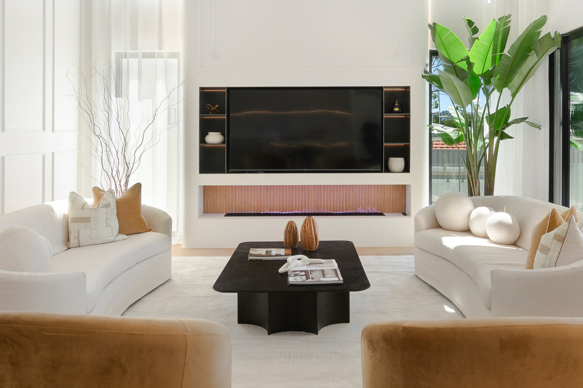 Close view of curved cream sofas, mustard velvet chairs and a black coffee table beneath a panelled wall and television