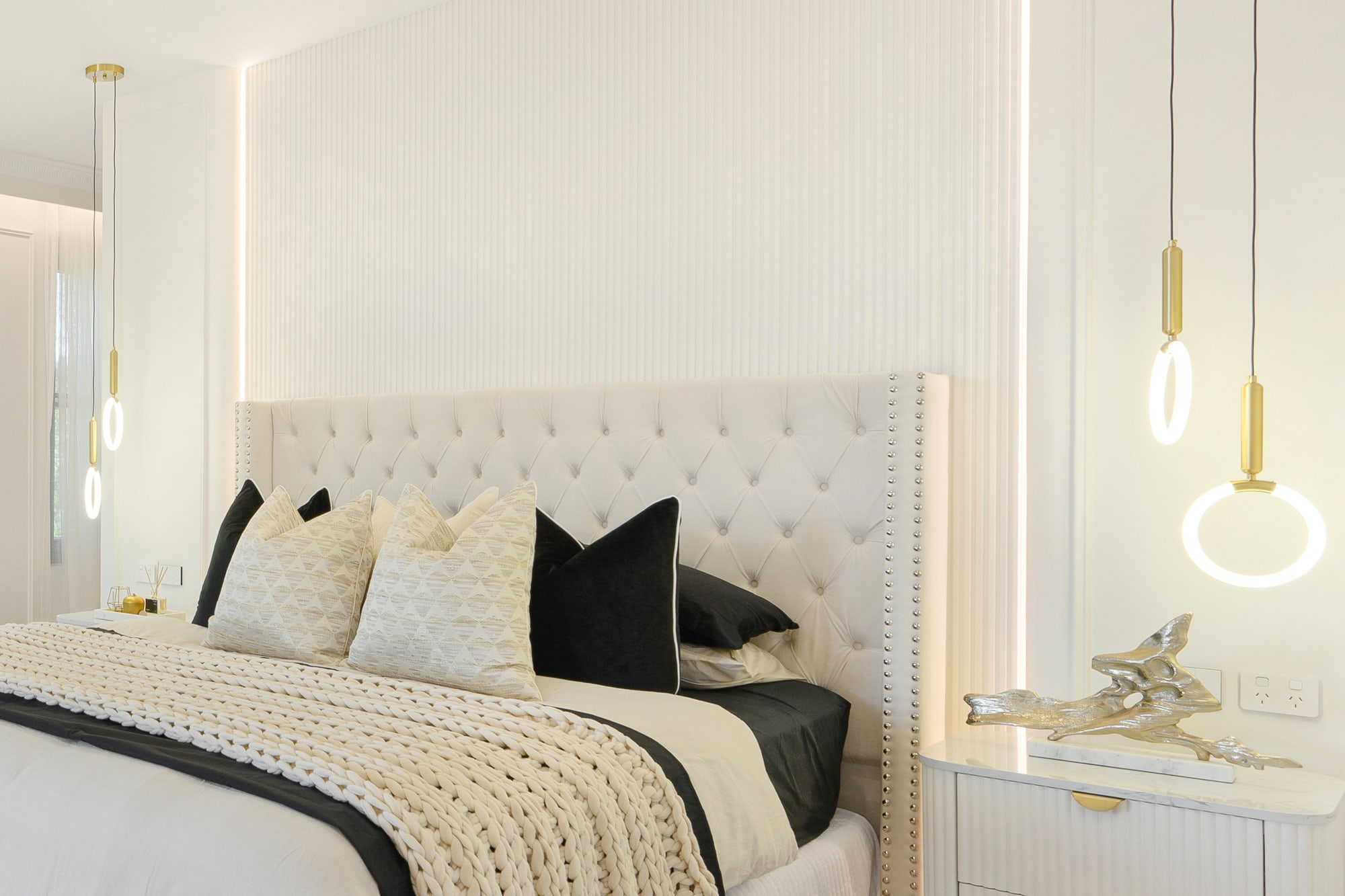 Tufted white bedhead with black and cream cushions, a knitted throw and gold pendant lights