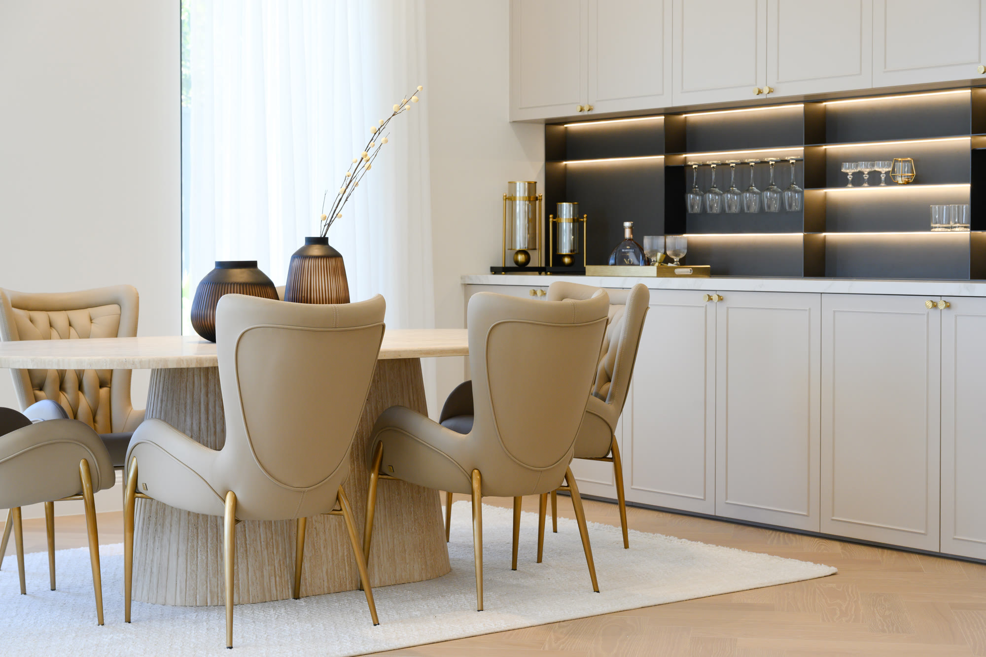 Dining setting with beige curved-back chairs and a round table in front of a bar-style cabinet with a glass shelf