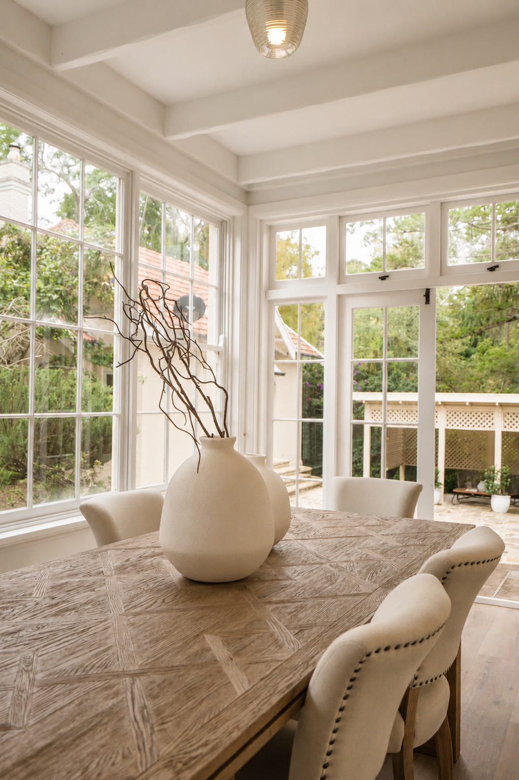 Dining table with cream studded chairs and a white vase beside a sunroom window
