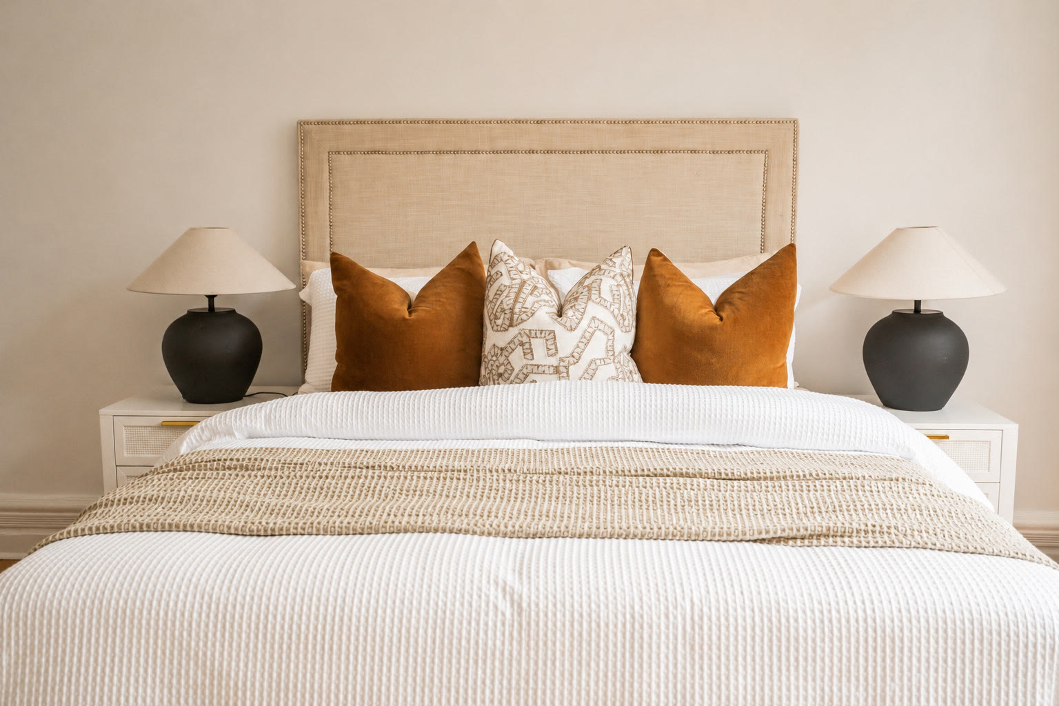 Bedroom with a linen bedhead, rust and patterned cushions and white bedside tables