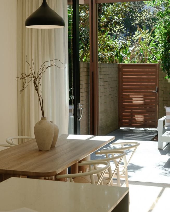 Dining table with a black pendant light and a vase of branches, with a courtyard seating area outside