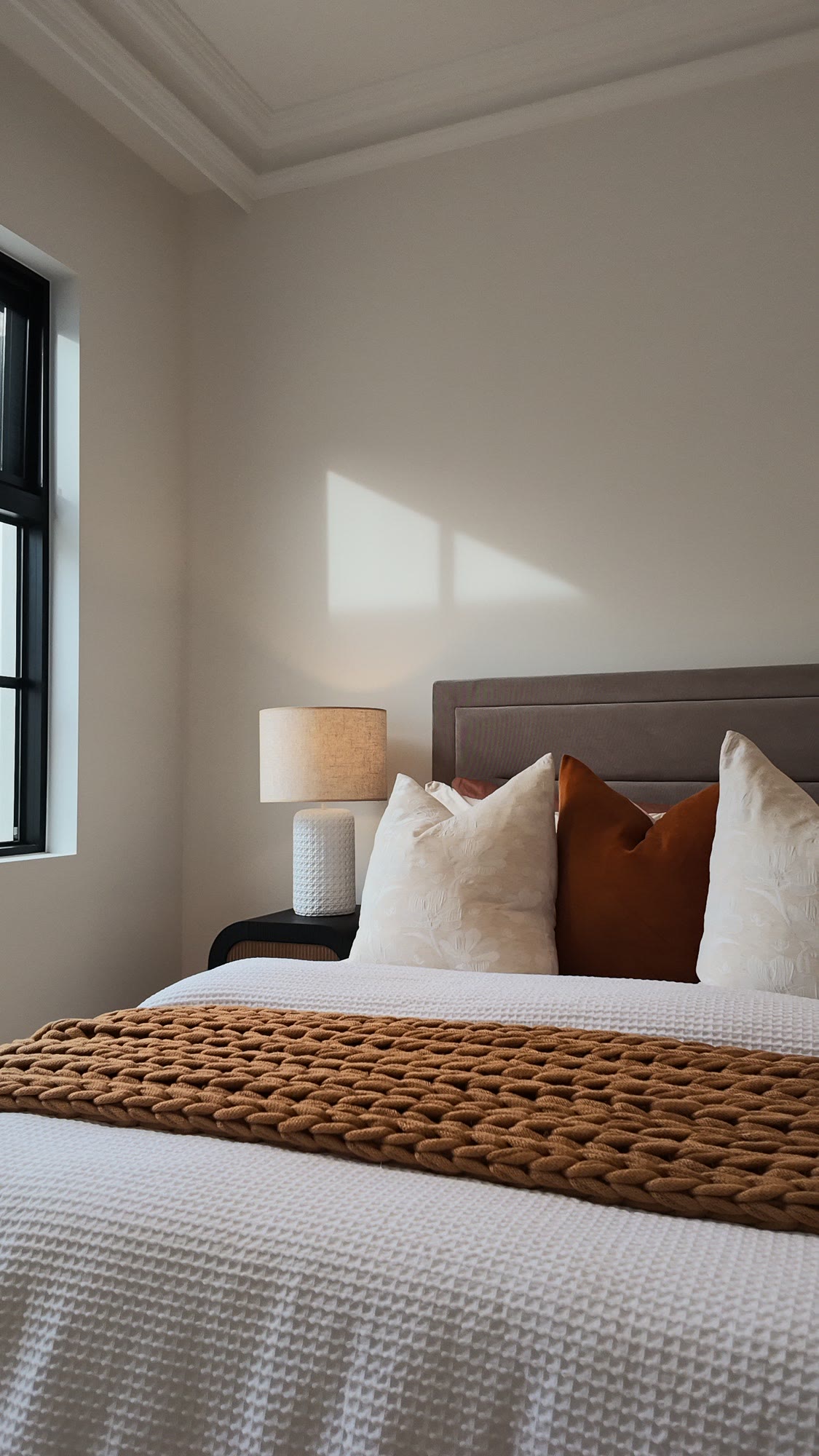 Bedroom with a rust knitted throw, cream and tan cushions and a black-framed window